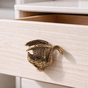 以下が含まれることがあります： 翼を広げた白鳥の形をした真鍮色の金属製引き出しノブ。明るい木目調の引き出しの前面に取り付けられています。白鳥は横向きで、羽の質感が細かく表現されており、首が右側に曲がったデザインになっています。