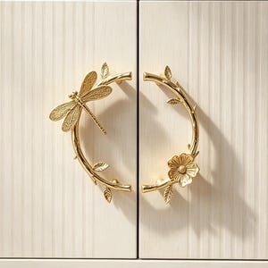 Könnte beinhalten: Ein weißer Schrank mit vertikal geriffelten Türen und zwei goldfarbenen Metallgriffen. Der linke Griff ist als gebogener Zweig mit einer Libelle geformt, der rechte Griff als gebogener Zweig mit einer Blume und kleinen Blättern.