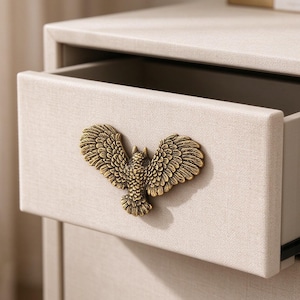 May include: A beige fabric-covered drawer featuring a brass-toned metal handle shaped like an owl with its wings spread wide. The drawer is partially open, revealing a smooth interior surface and a clean, rectangular design on the front panel.