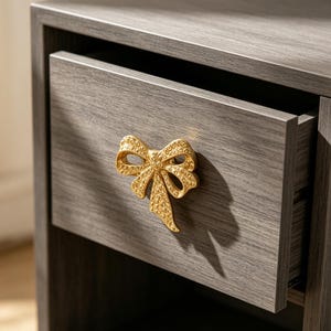 May include: A grey wood-grain nightstand drawer featuring a metallic gold bow-shaped handle. The handle has a textured, dimpled surface and is centred on the front panel of the drawer, which is partially pulled open to reveal the interior.