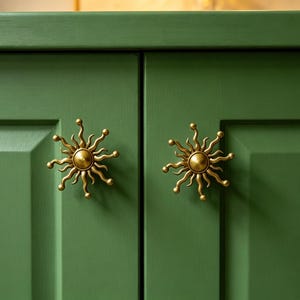 May include: Two brass-colored metal cabinet knobs shaped like suns with radiating wavy rays, mounted on a pair of matte forest green cabinet doors. Each knob features a central dome with multiple curved, pointed arms extending outward.