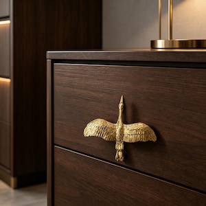 Puede incluir: Un frente de cajón de madera marrón oscuro con un tirador de mueble de color dorado metálico con forma de ave con las alas completamente extendidas en vuelo. El tirador está centrado en el cajón y muestra texturas detalladas de plumas.