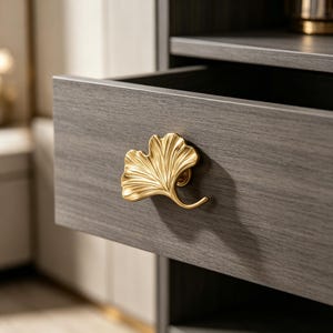 Puede incluir: Un tirador de cajón de metal dorado con forma de hoja de ginkgo fijado a un cajón de madera gris oscuro con vetas. El diseño de la hoja presenta nervaduras detalladas y un tallo curvado, sirviendo como manija funcional para el cajón.