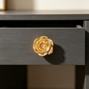 Könnte beinhalten: Ein goldfarbener Schubladengriff aus Metall in Form einer blühenden Blume mit geschichteten, strukturierten Blütenblättern. Der Griff ist an der Vorderseite einer dunkelgrauen Schublade mit Holzmaserung befestigt und bildet einen Kontrast.