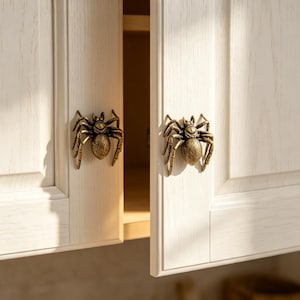 May include: Two antique brass-colored metal spider-shaped cabinet knobs attached to light-colored wood-grain cabinet doors. Each knob features a detailed eight-legged spider design with a textured body and curved legs, serving as a functional handle.