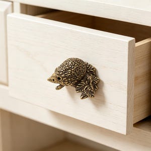May include: A brass-toned metal drawer knob shaped like a hedgehog resting on a cluster of leaves. The detailed, textured animal figure is mounted onto the front of a light-coloured, natural wood drawer with a visible grain pattern and finish.