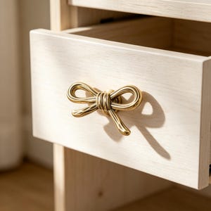 May include: A light-coloured wooden drawer featuring a polished brass cabinet knob shaped like a decorative bow. The bow-shaped handle is centred on the drawer front, which displays a visible wood grain texture and a smooth, matte finish.