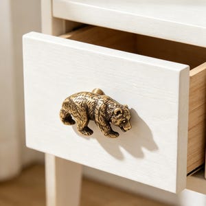 Könnte beinhalten: Ein schrankknopf aus messingfarbenem metall in form eines gehenden bären, montiert an einer weißen schubladenfront aus holz. Die bärenfigur weist eine detaillierte fellstruktur und ein metallisches finish auf und ist mittig auf der platte.