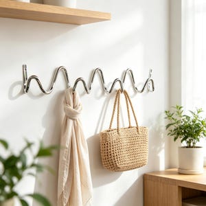 Peut inclure: Un porte-manteau mural en métal chromé poli au design ondulé et sinueux continu. Le support comporte plusieurs crochets pour suspendre des objets, avec une écharpe en tissu beige et un sac fourre-tout en paille tressée suspendus aux crochets.