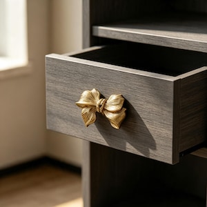 May include: A dark gray wood-grain drawer featuring a metallic gold-toned bow-shaped handle. The drawer is partially pulled open, revealing a hollow interior space, with the bow handle centered on the front panel of the drawer unit.