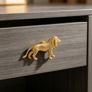 May include: A gold-colored metal drawer pull shaped like a standing lion with a detailed mane and tail. The lion figure is mounted horizontally on the front of a dark gray wood-grain drawer, serving as a functional handle for opening the furniture.