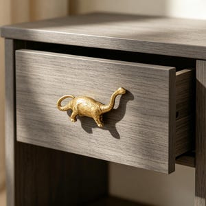 May include: A gray wood-grain drawer front featuring a metallic gold-toned dinosaur-shaped handle. The drawer is slightly open, revealing the interior side panel. The dinosaur figure is depicted in profile, facing right, with a long neck and tail.