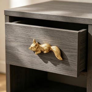 May include: A dark gray wood grain nightstand featuring a single drawer with a gold-toned metal fox-shaped handle. The fox is depicted in a horizontal, running position with detailed fur texture and a long, curved tail extending to the right side.
