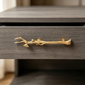 May include: A gold-coloured metal drawer pull shaped like a textured tree branch with small twig details. The handle is mounted horizontally onto a dark grey wood-grain drawer front, featuring two attachment points that secure it to the surface.