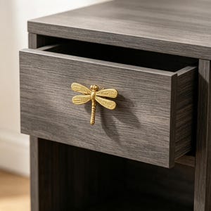 May include: A dark grey wood-grain nightstand featuring a single pull-out drawer. The drawer is accented with a prominent, textured gold-toned metal dragonfly-shaped handle centred on the front panel, providing a distinct contrast to the wood.