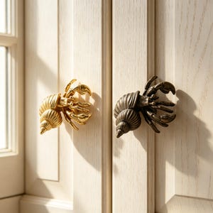 May include: Two cabinet knobs shaped like hermit crabs, one in a polished gold finish and the other in an antique bronze finish. Each knob features a detailed spiral shell and crab legs, mounted on light-colored wooden cabinet doors.