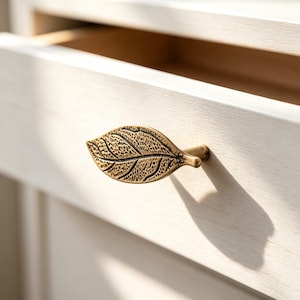 May include: A brass-toned metal drawer knob shaped like a leaf with a textured surface and detailed veins. The knob is attached to a light-colored wooden drawer front, casting a distinct shadow on the surface of the wood to the right side.