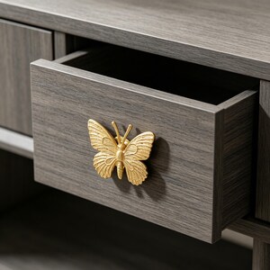 May include: A gold-coloured metal butterfly-shaped drawer pull attached to a dark grey wood-grain drawer. The drawer is partially open, revealing the interior space. The butterfly handle features detailed wing textures and two thin antennae.