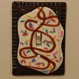 May include: A rectangular light switch cover plate designed to resemble a pastry with a dark brown rectangular base, a white icing layer, brown icing drizzle, and colorful sprinkles. The switch toggle is white and positioned in the center.