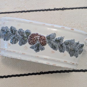 Può includere: Un piatto da portata ovale in ceramica con smalto color crema e un motivo decorativo in rilievo che presenta una pigna marrone centrale fiancheggiata da foglie verdi testurizzate. Il piatto include piccole maniglie su ogni lato e sottili tocchi.