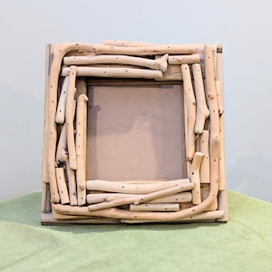 May include: A square picture frame constructed from light-colored, smooth driftwood pieces arranged in a layered, overlapping pattern around a central rectangular opening with a plain, neutral-toned backing board visible inside.