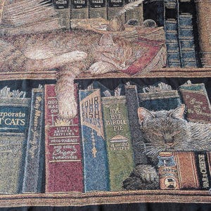 May include: A woven throw blanket featuring a library scene with two sleeping cats. The design includes shelves filled with books titled Cat Tales, Corporate Cats, Four Star Fish, Bye Bye Birdie Pie, and War & Cheese, along with a quill and inkwell.
