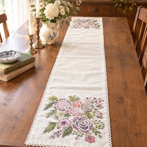 Puede incluir: Un camino de mesa rectangular de color blanco roto con intrincados bordados florales en tonos morados, rosas y verdes en ambos extremos. El camino está rematado con un delicado borde de encaje de ganchillo blanco en todo su contorno.