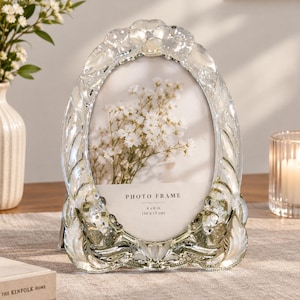 May include: An oval-shaped clear glass photo frame with a decorative floral relief border. The frame features a central insert with the text "PHOTO FRAME" and "4 x 6 in (10 x 15 cm)" printed on it, showcasing a white floral graphic background.