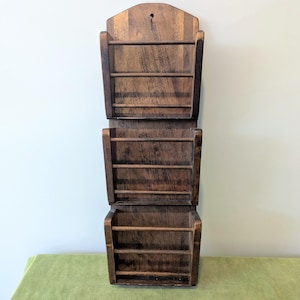 May include: A vertical wall-mounted wooden organiser featuring three stacked storage compartments. Each compartment has two horizontal wooden dowels across the front to secure items. The unit is constructed from dark brown stained wood with a visible grain.