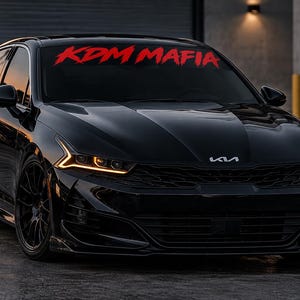 May include: A black Kia sedan featuring a red KDM MAFIA decal across the top of the windshield. The vehicle has dark tinted windows, black multi-spoke wheels, and amber LED daytime running lights that illuminate the front grille and headlights.