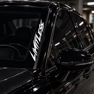 May include: A white vinyl decal applied to the passenger side of a black car windshield. The decal features the word LIMITLESS written in a sharp, jagged, stylized font oriented vertically along the edge of the glass near the A-pillar.