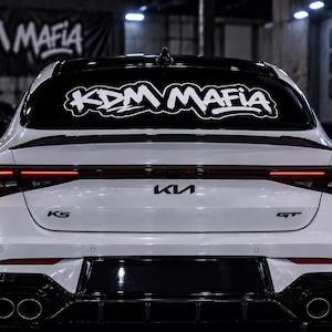 May include: A white Kia K5 GT sedan viewed from the rear, featuring a black roof, a black rear spoiler, and a quad-tip exhaust system. The rear window displays a large white decal with the text KDM MAFIA in a stylized, bold, graffiti-inspired font.