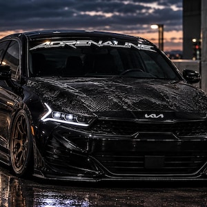 May include: A black Kia sedan with a carbon fiber hood, bronze multi-spoke wheels, and sharp LED headlights. The windshield features a white decal with the text ADRENALINE. The vehicle has a lowered stance and a glossy finish reflecting ambient light.