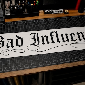 May include: A long white vinyl decal featuring the text Bad Influence in a bold black gothic font. The lettering includes decorative flourishes and swirls extending from the bottom of the characters, creating a stylized script appearance.