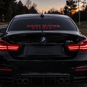 May include: A black BMW M4 sports car viewed from the rear, featuring bright red illuminated taillights, a quad exhaust system, and a red decal on the rear window displaying the text NIGHT RUNNER in English and Japanese katakana characters.