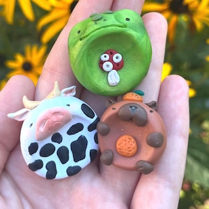 May include: Three small, round, sculpted clay figures. One is a bright green frog with a red and white mushroom on its belly. Another is a white cow with black spots and small horns. The third is a brown capybara with a small orange on its head.