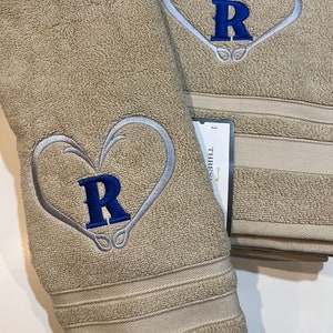 May include: Two beige bath towels with embroidered fishing hooks forming a heart shape with the letter "R" in blue inside. The towels have a THRES... label.