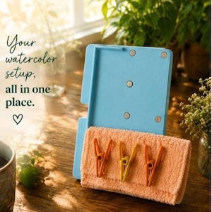 May include: A light blue rectangular board with four circular magnets, holding a folded peach-colored towel secured by three plastic clips. The text reads: Your watercolor setup, all in one place.