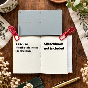 May include: A rectangular light gray metal tool with four circular magnetic points, positioned above an open sketchbook held by two red binder clips. The sketchbook pages feature the text 5.35x3.86 sketchbook shown for reference and Sketchbook not included.