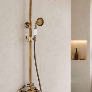 Könnte beinhalten: Ein wandmontiertes Duschsystem in antikem Messing-Finish mit einem runden Regenduschkopf, einer Handbrause mit weißem Keramikgriff und einer Armatur mit zwei weißen Keramik-Hebelgriffen. Der Text lautet BrassTap.