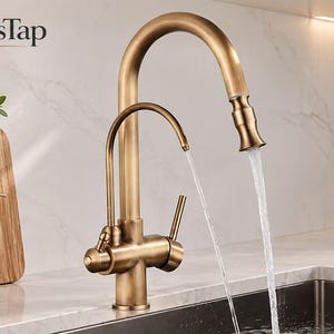 May include: A brushed brass kitchen faucet with a high-arc pull-down sprayer and a secondary smaller water spout. The faucet features a single lever handle and is dispensing water from both outlets. The text BrassTap is visible in the top left corner.