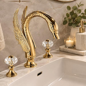 May include: A gold-coloured swan-shaped tap with textured wings and two clear, faceted crystal handles. The tap features the text BrassTap at the top left. It is mounted on a white marble countertop next to a white sink basin.
