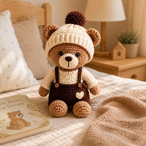 Peut inclure: Un ours en peluche crocheté portant un bonnet crème avec un pompon marron foncé, un haut à manches longues crème et une salopette marron foncé avec deux petits boutons et un petit écusson en forme de cœur crème sur le devant.