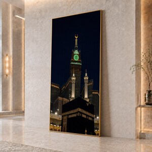 May include: A vertical rectangular wall art piece featuring a night scene of the Kaaba in Mecca with the illuminated Abraj Al-Bait clock tower in the background. The image is framed in a thin gold border and displays the black cube structure.