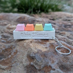 May include: A white rectangular keychain base featuring the embossed text Ohio Slimes on the front. Four small, pastel-colored square keycaps in pink, orange, yellow, and light blue are mounted on top of the white base, with a silver metal ring attached.