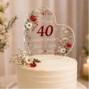 May include: A heart-shaped clear acrylic cake topper featuring a floral design with red poppies and white daisies. The text reads Happy 40th ANNIVERSARY James & Linda 1986 2026. The topper is decorated with small white and red flowers at the base.