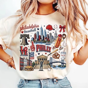 May include: A cream-colored t-shirt featuring a graphic print of Philadelphia landmarks, a hot dog mascot, the Liberty Bell, a baseball, a bat, a pretzel, and the text "philadelphia baseball" and "PHILLY" with red, blue, and brown accents.