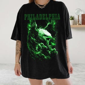 May include: A black short-sleeve t-shirt featuring the word PHILADELPHIA in green block letters at the top. Below the text is a graphic of a bald eagle with green glowing feathers and lightning effects, depicted with its wings spread and talons out.
