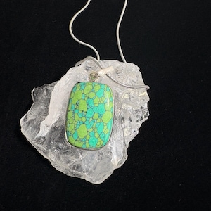 May include: A rectangular pendant featuring a vibrant green and turquoise mosaic pattern with dark veining, set in a silver-toned metal frame. The pendant hangs from a thin, flexible silver-toned snake chain, resting upon a clear, jagged crystal.