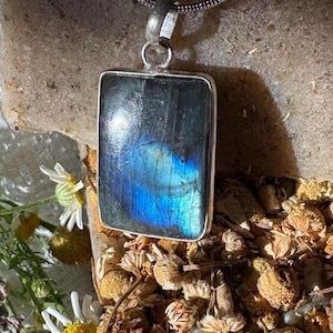 May include: A rectangular labradorite pendant set in a silver-toned metal frame. The stone displays a dark grey base color with a prominent, iridescent blue flash across its center. The pendant is attached to a silver-toned chain bail at the top.